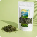 Loser Sencha Grüntee von MEE KAFFEE mit sorgfältig verarbeiteten, dunkelgrünen Teeblättern neben der Verpackung. Der traditionelle japanische Grüntee überzeugt durch sein frisches Aroma, feine Umami-Noten und eine schonende Verarbeitung für höchsten Teegenuss.